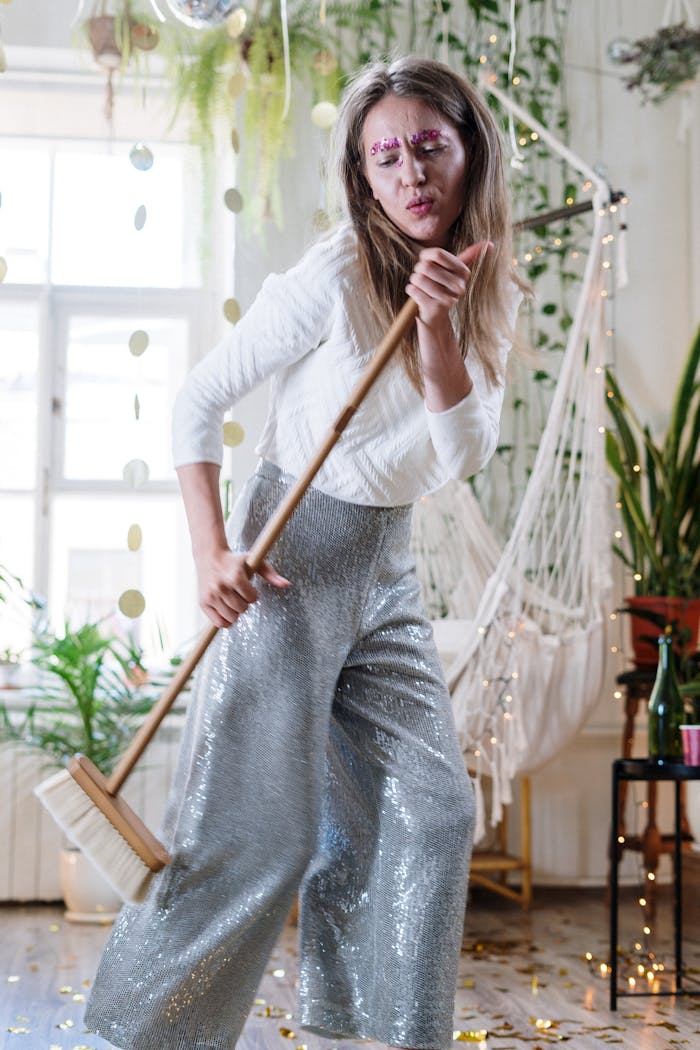 Woman in sparkly pants joyfully cleaning up with a broom after a festive party indoors.